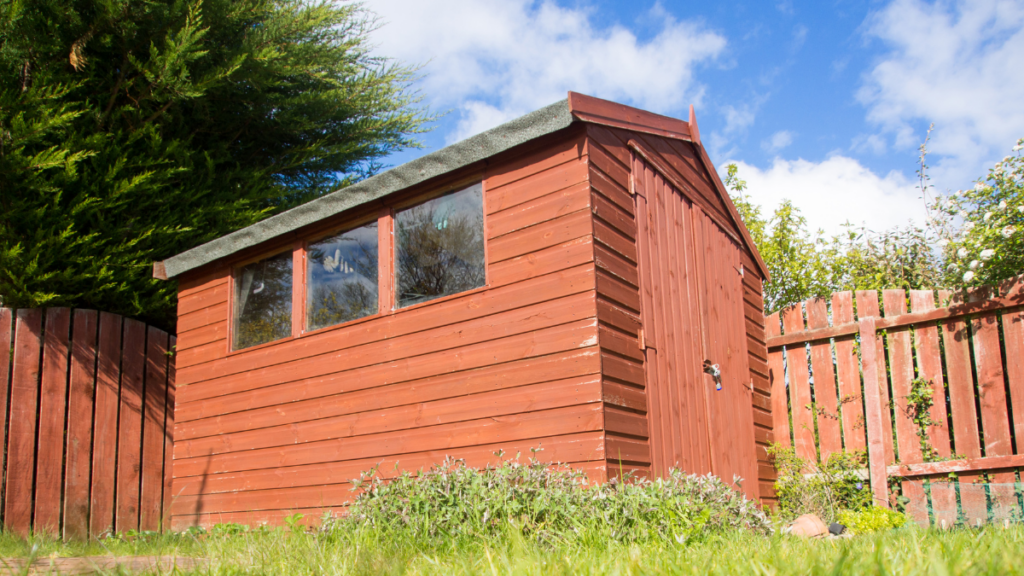 Wooden garden shed painted in warm reddish-brown, complemented by a backdrop of lush green foliage and set against a clear blue sky.