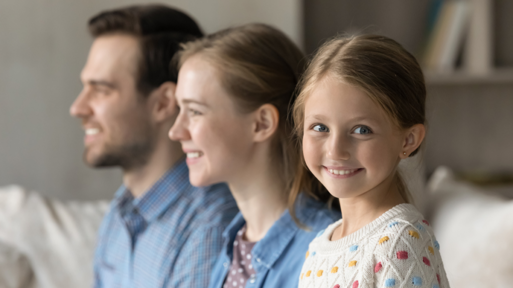 Side profile of a happy family seated together, with a young girl in the foreground smiling at the camera, embodying warmth and togetherness.