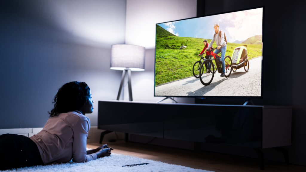Cozy home scene of a woman lying on a soft rug, enjoying a vibrant movie on a modern flat-screen TV, with ambient lighting from a floor lamp in the background.