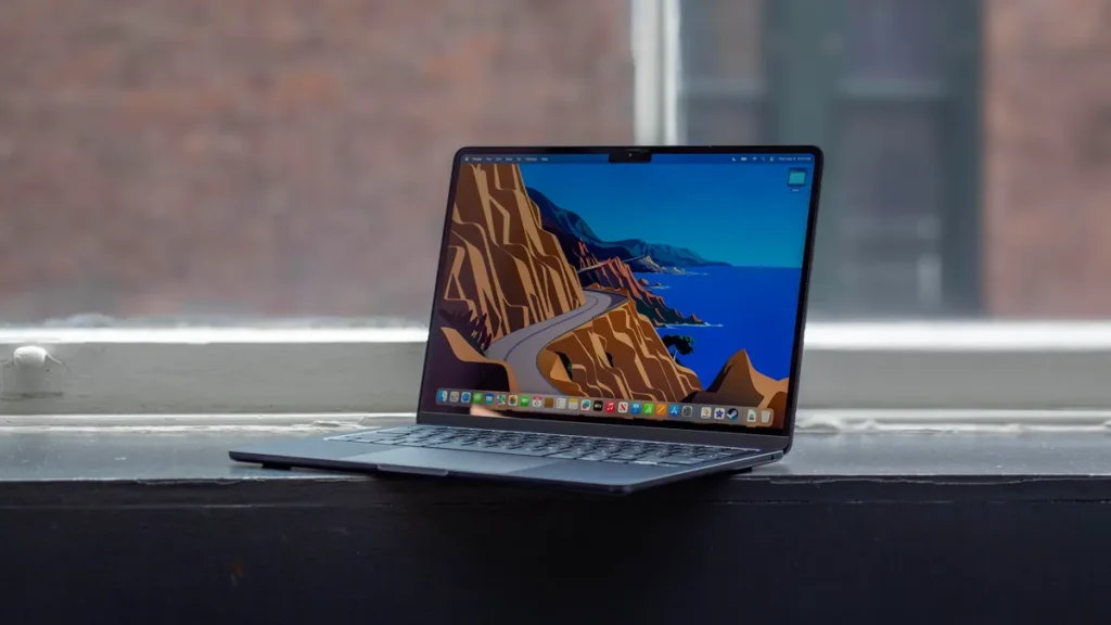 Laptop placed on a windowsill, displaying a vivid screen with a scenic digital wallpaper featuring a winding road and mountains.