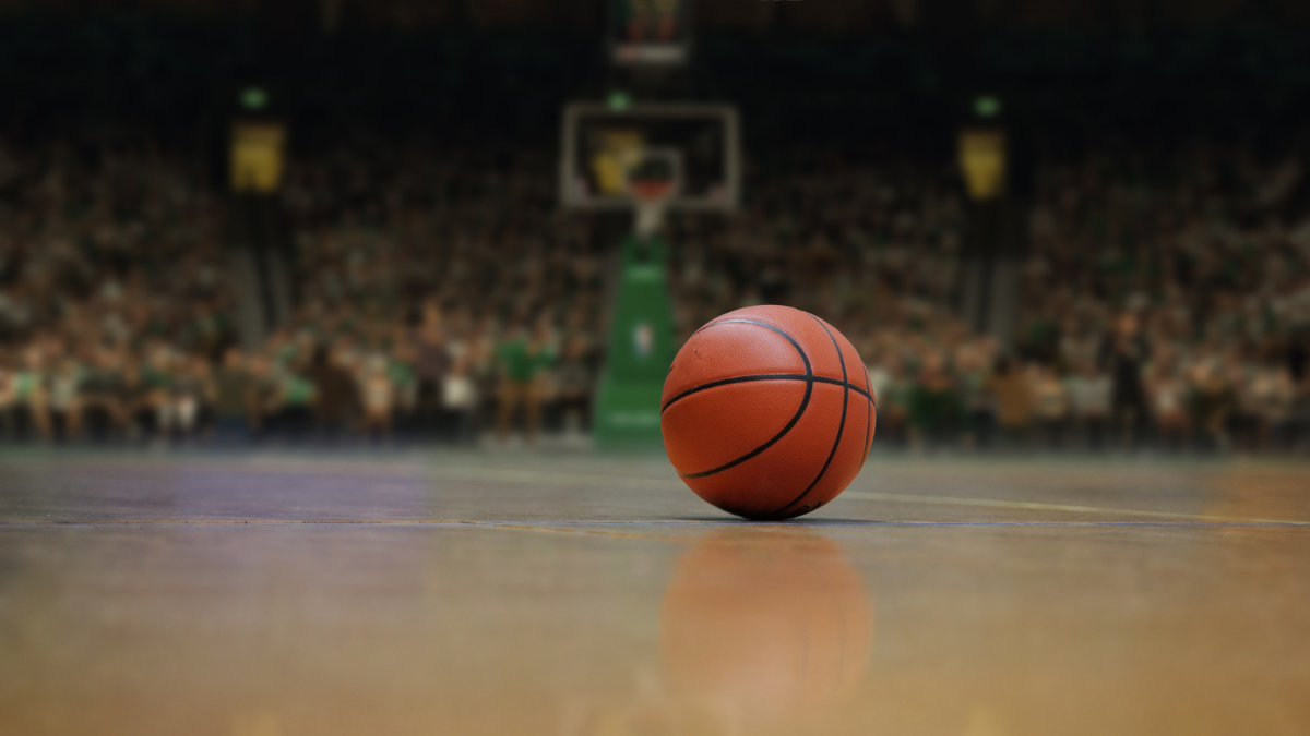A basketball resting on the court floor with a blurred crowd and hoop in the background, symbolizing anticipation before the game.