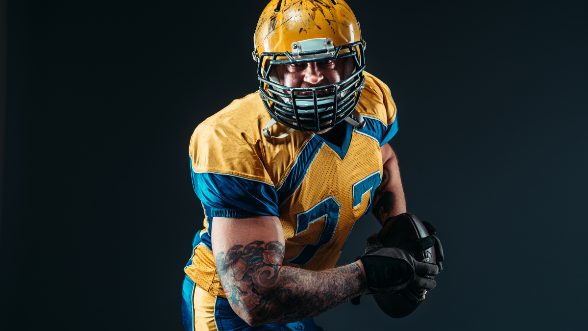 A focused football player in yellow and blue gear, gripping the ball, ready for action under dramatic lighting.