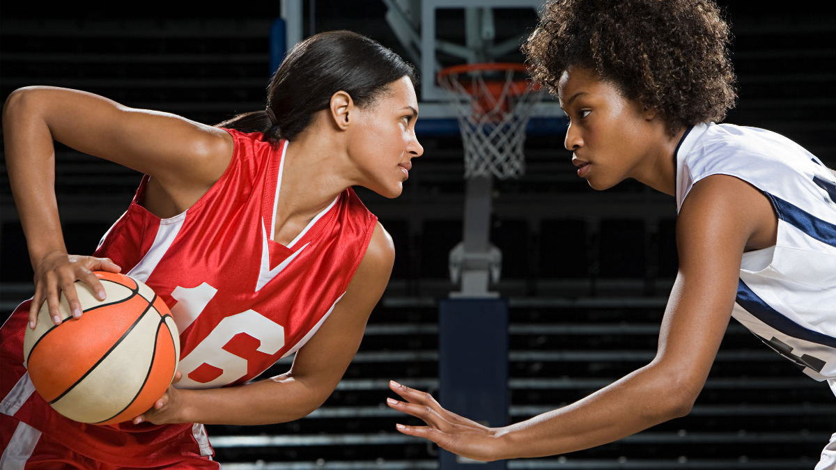 Two female basketball players in an intense one-on-one moment, one in red holding the ball, and the other in white defending, with a hoop visible in the background.