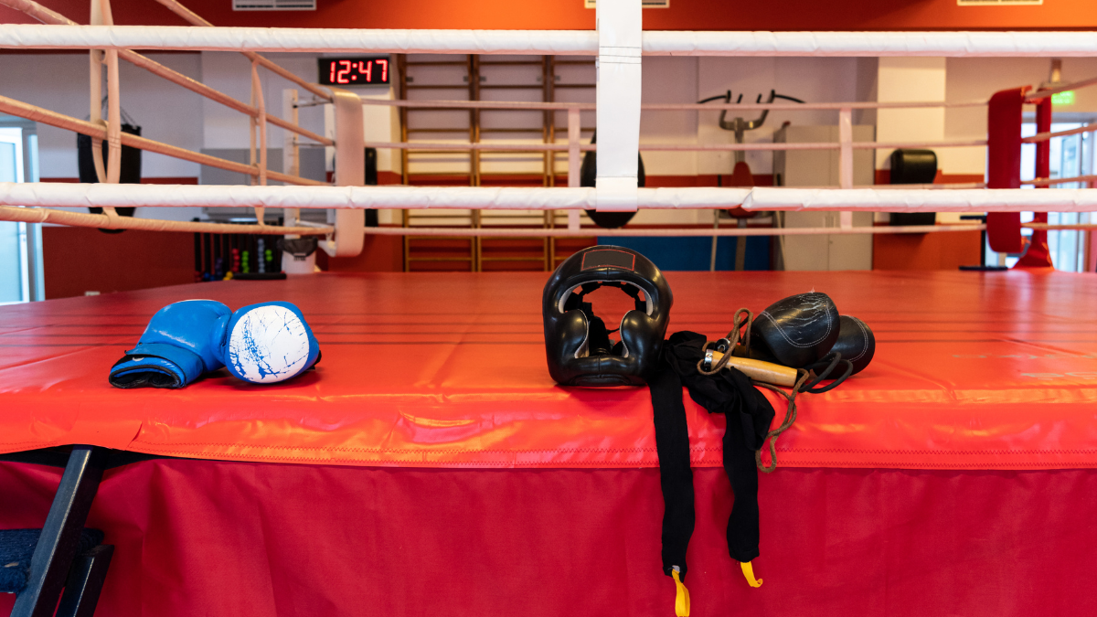 A vibrant boxing ring setup featuring red padding, gloves, and protective gear draped over the ropes, with a digital clock and gym equipment visible in the background.