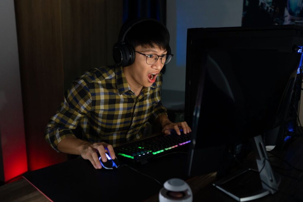 Smiling gamer wearing a headset and cap, streaming at a setup with dual monitors, a ring light, and colorful LED backlighting.