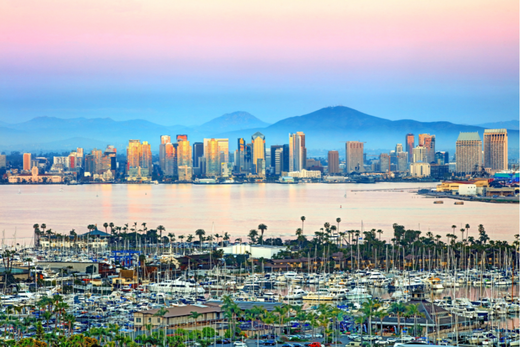 Scenic view of San Diego's downtown skyline at sunset, with boats docked in the marina and mountains in the background.