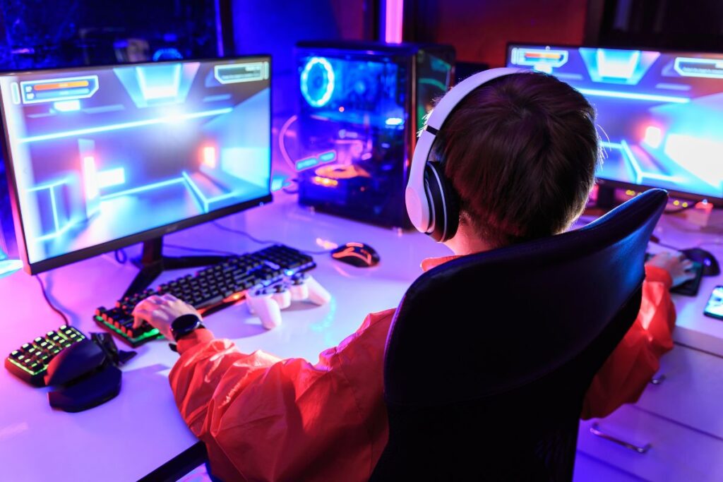 A focused gamer holds a controller while playing on a monitor, illuminated by purple and blue ambient lighting.