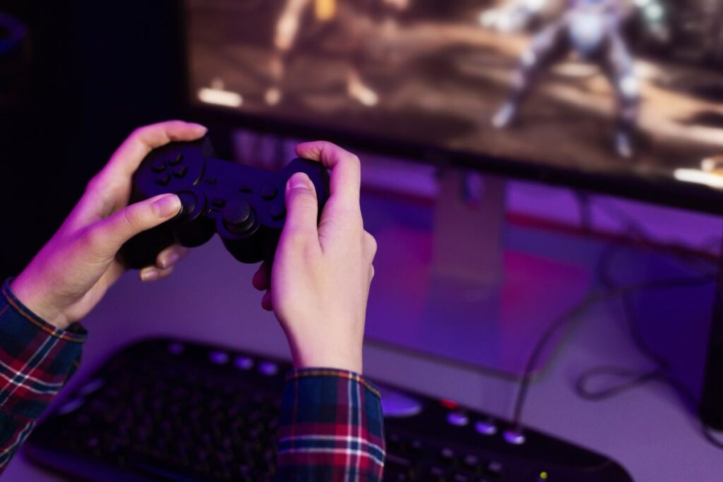 Close-up of hands holding a game controller in front of a screen, immersed in gameplay with a dimly lit gaming setup.