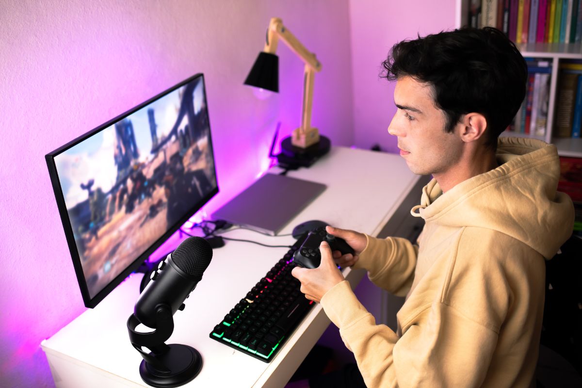 A focused gamer wearing a hoodie plays with a controller at a desk setup, with a microphone and purple LED-lit monitor.