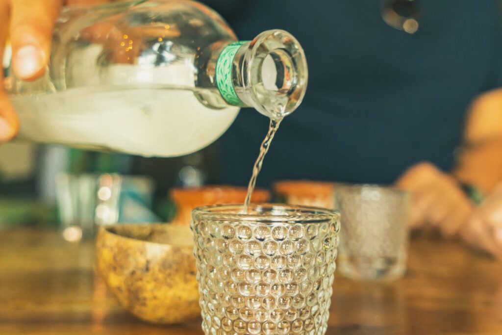 Hand pouring clear liquid into a textured glass on a table.