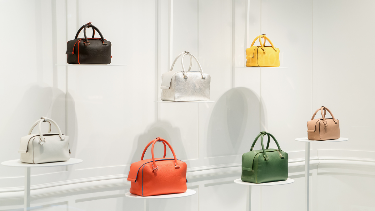 Display of six colorful handbags in a minimalist boutique setting, featuring shades of black, white, yellow, red, green, and beige on elevated stands.