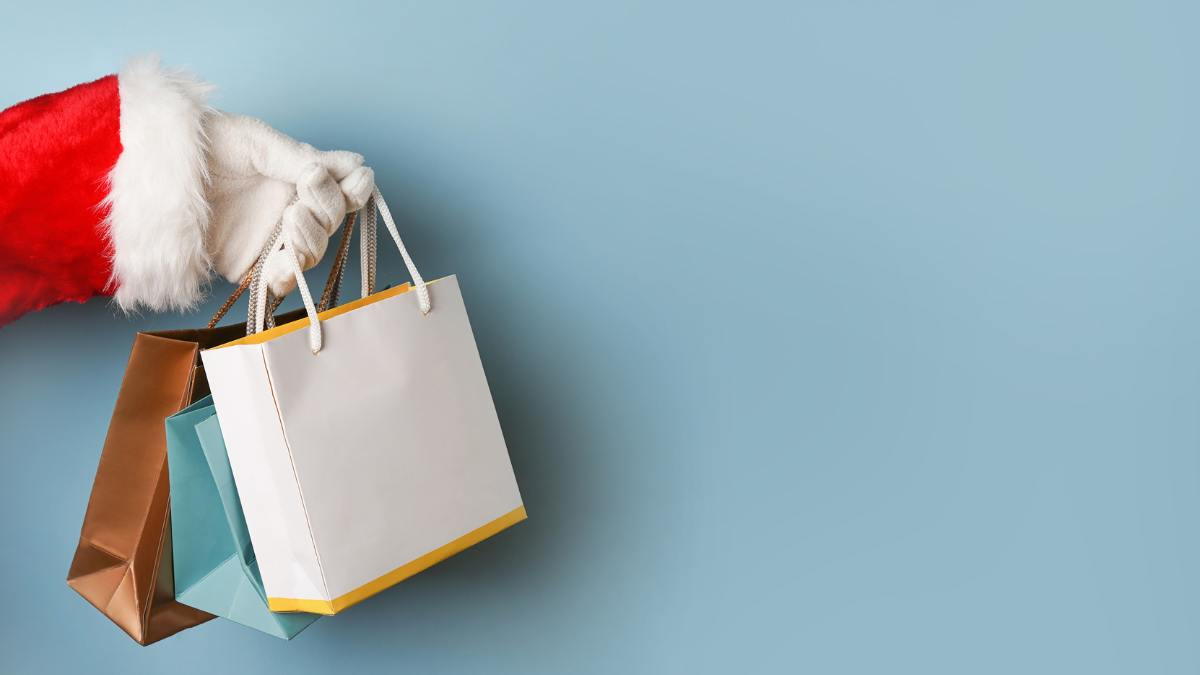 Santa Claus hand holding festive shopping bags against a light blue background, symbolizing holiday shopping and gift-giving season.