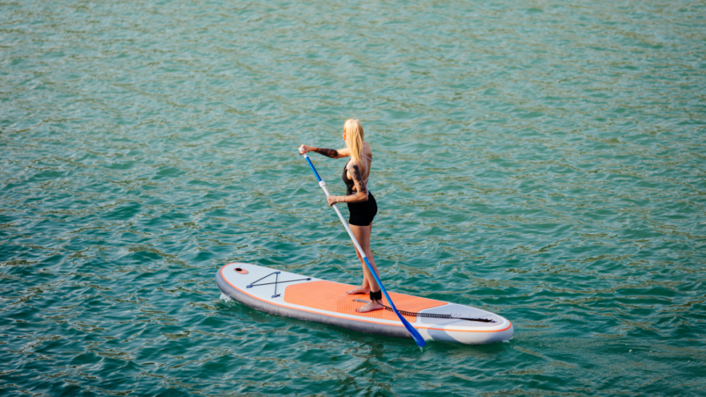 Person paddleboarding on a calm body of water using an orange and white stand-up paddleboard  and a blue paddle. The individual maintains balance while standing and propelling forward.