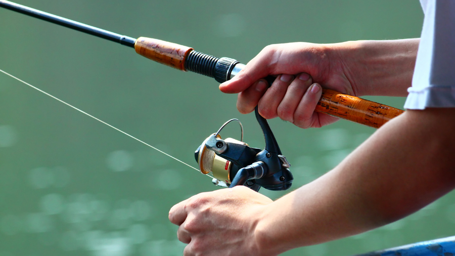 A close-up of a fisherman’s hands gripping a spinning reel fishing rod over a calm body of water.