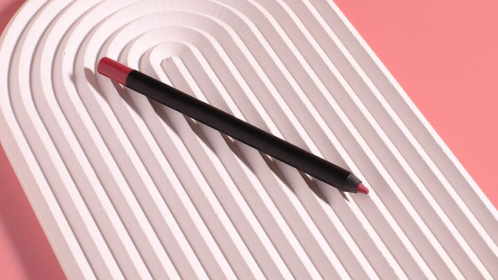 A black lip liner pencil with a pink tip is placed on a textured white tray with a pink background.