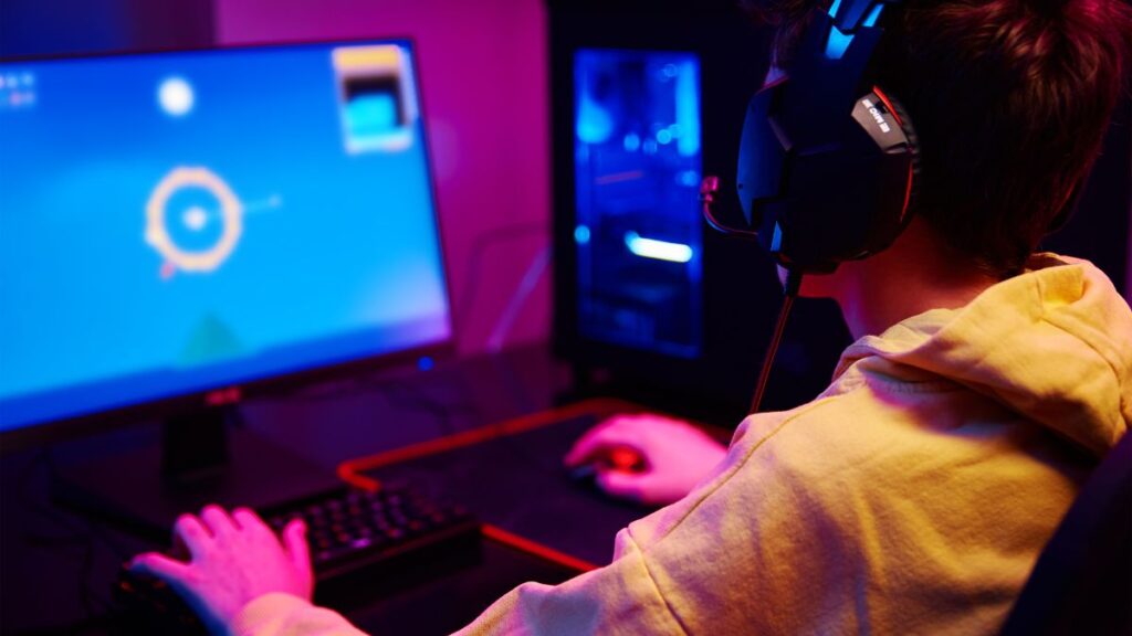 A person wearing a headset and a yellow hoodie is playing a computer game in a dimly lit room with blue and purple lighting.