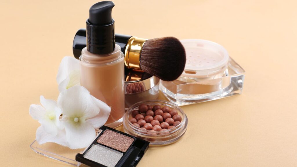 A bottle of liquid foundation with a makeup brush, loose powder, bronzing pearls, and eyeshadow on a beige background.