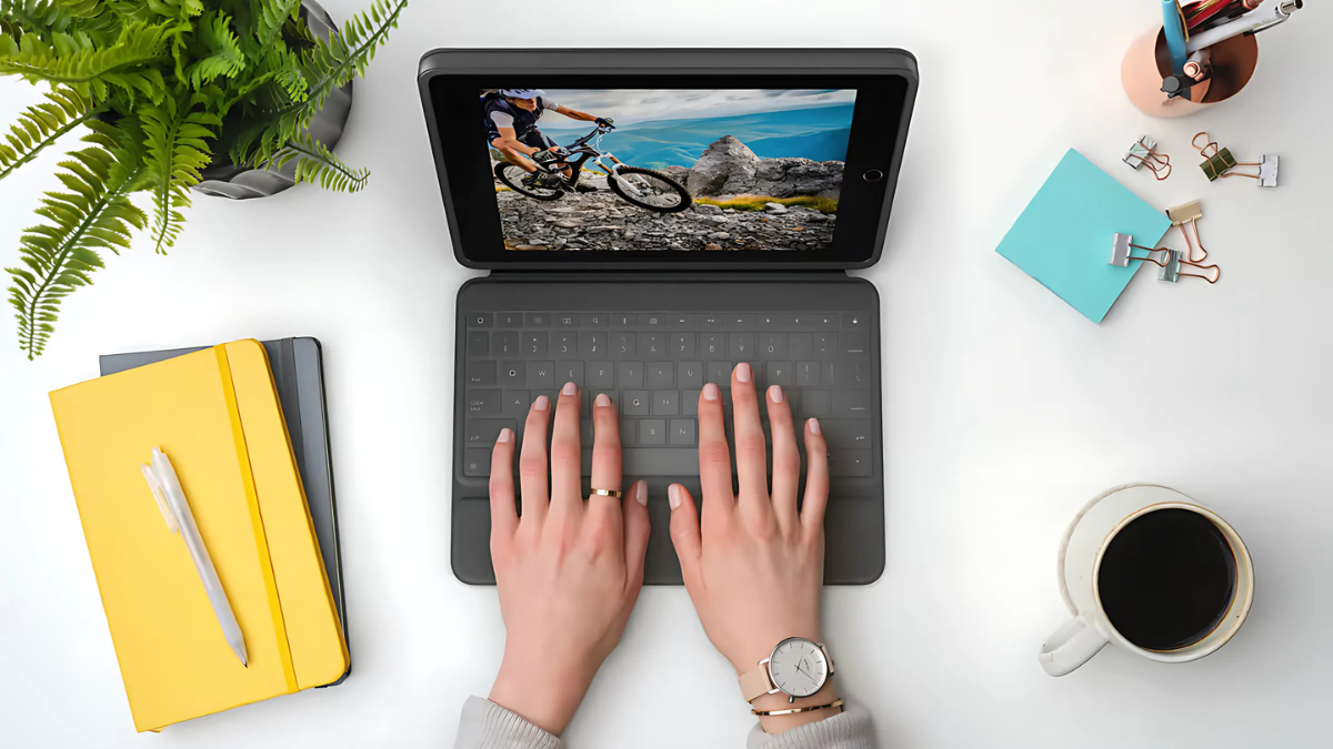 Top view of a person typing on a tablet with a detachable keyboard, surrounded by coffee, notebooks, and office supplies