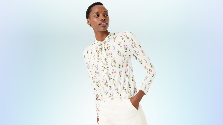 Woman wearing a white long-sleeve floral blouse tucked into white pants, posing confidently against a light gradient background.