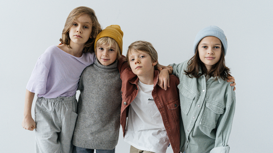 Four stylish children standing together with arms around each other, dressed in casual, cozy outfits with beanies.