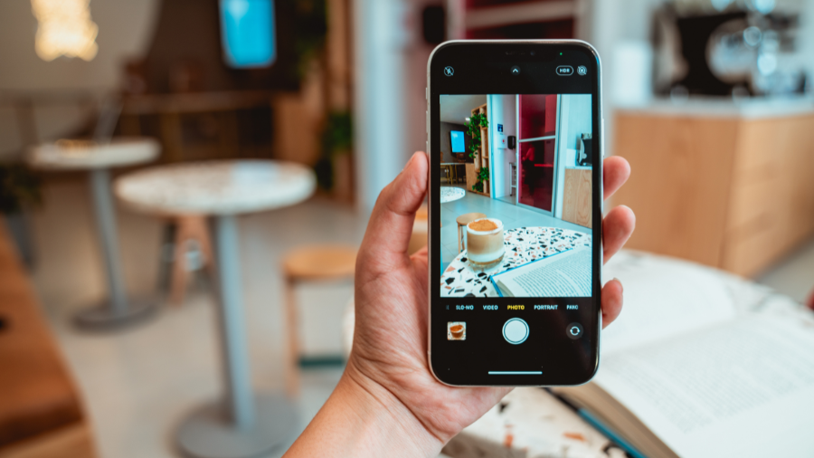 Hand holding a smartphone taking a photo of a coffee cup on a table in a cozy café setting.