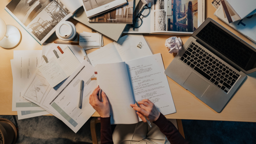 Person studying at a desk with open books, papers, a laptop, and a coffee cup.