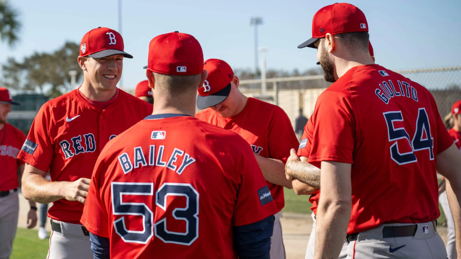 Boston Red Sox players in red jerseys and matching caps gather on a sunny day during spring training, sharing a conversation and smiles on the field.