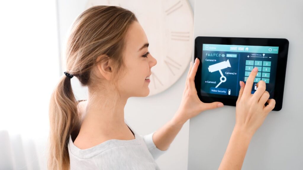 A woman is smiling while using a wall-mounted smart home security system on a touchscreen panel.
