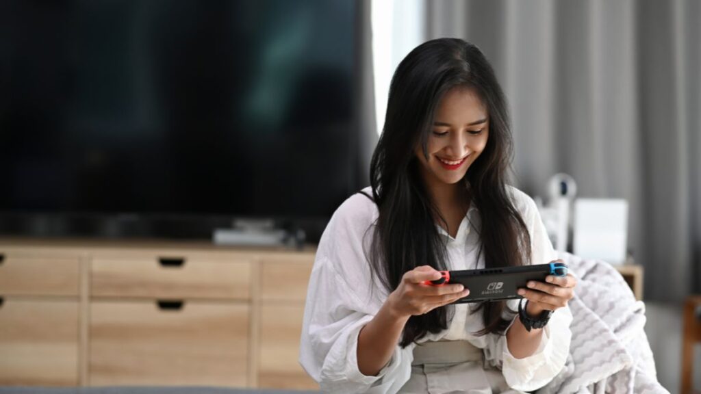 A smiling woman in a white outfit sits on a couch playing a Nintendo Switch.