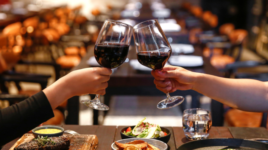 Two people clinking red wine glasses over a dinner table at a restaurant.