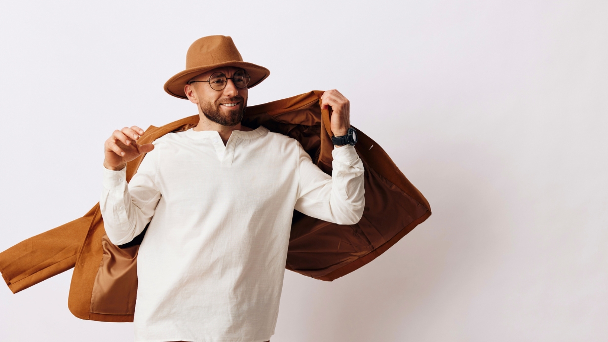 Stylish man wearing a brown hat, white shirt, and brown coat posing confidently against a light background — modern casual fashion look for men